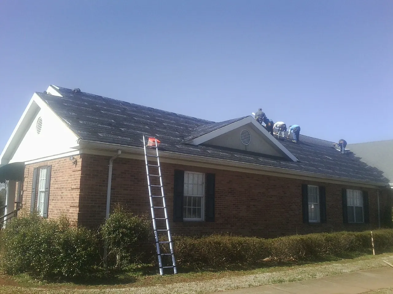Crew tearing shingles off a brick ranch roof with a ladder set — Metal Roof Repair work by our crew in Whiteville