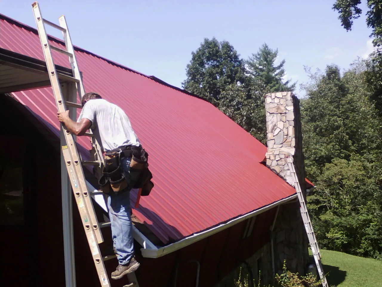 Roofer on a ladder beside a red metal roof and stone chimney — Soffit & Fascia Repair by Roofing Brothers in Whiteville